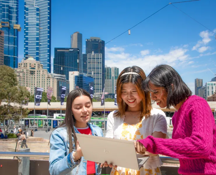 Three female international students looking at laptop on balcony overlooking the city